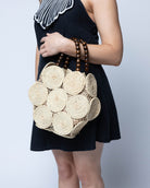 Model wearing Romy Natural Crochet Disc Straw Handbag with Wooden Bead Handles handmade natural raffia palm fiber in a solid hue of natural straw color  in a crochet coiled circular flat disc attached together with macrame knot technique with a geometric hexagonal overall shape with seven discs per side two above and below a horizontal row of three per side and a single row comprising the side of the bag with dark brown carved round wooden beaded handles purse - Shebobo