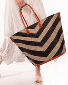 Model wearing Santa Cruz Wide Stripe Chevron Large Straw Tote Bag handmade loomed raffia palm fiber in a two tone diagonal even width bands of color in a wide stripe chevron pattern of black and natural straw color on the front and a matching solid hue on the back with leather accents and handles large shoulder bag - Shebobo