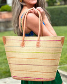 Model wearing Santa Cruz Brights Pinstripes Small Straw Tote Bag handmade loomed raffia in a wide stripe of natural straw color with multicolor brights thin stripes of fuchsia pink, grey, lime green, bordeaux deep red, coral orange, tea brown, etc. in a horizontal pinstripe pattern handbag with leather handles and details beach bag - Shebobo