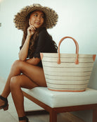 Model wearing Santa Cruz Natural Pinstripes Small Straw Tote Bag handmade loomed raffia in a horizontal pinstripe pattern of wide bands of natural straw color with narrow black bands handbag with leather handles and accents - Shebobo (with 5" wide brim Kat Black Two Tone Multicolor Straw Sun Hat with Raw Fringe Edge)