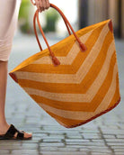Model wearing Santa Cruz Wide Stripe Chevron Large Straw Tote Bag handmade loomed raffia palm fiber in a two tone diagonal even width bands of color in a wide stripe chevron pattern of saffron orange/yellow and natural straw color on the front and a matching solid hue on the back with leather accents and handles large shoulder bag - Shebobo