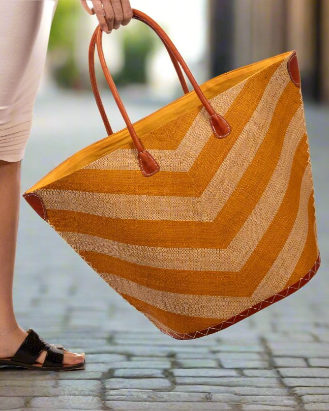 Model wearing Santa Cruz Wide Stripe Chevron Large Straw Tote Bag handmade loomed raffia palm fiber in a two tone diagonal even width bands of color in a wide stripe chevron pattern of saffron orange/yellow and natural straw color on the front and a matching solid hue on the back with leather accents and handles large shoulder bag - Shebobo