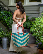 Model wearing Santa Cruz Wide Stripe Chevron Large Straw Tote Bag handmade loomed raffia palm fiber in a two tone diagonal even width bands of color in a wide stripe chevron pattern of teal green/blue and natural straw color on the front and a matching solid hue on the back with leather accents and handles large shoulder bag - Shebobo