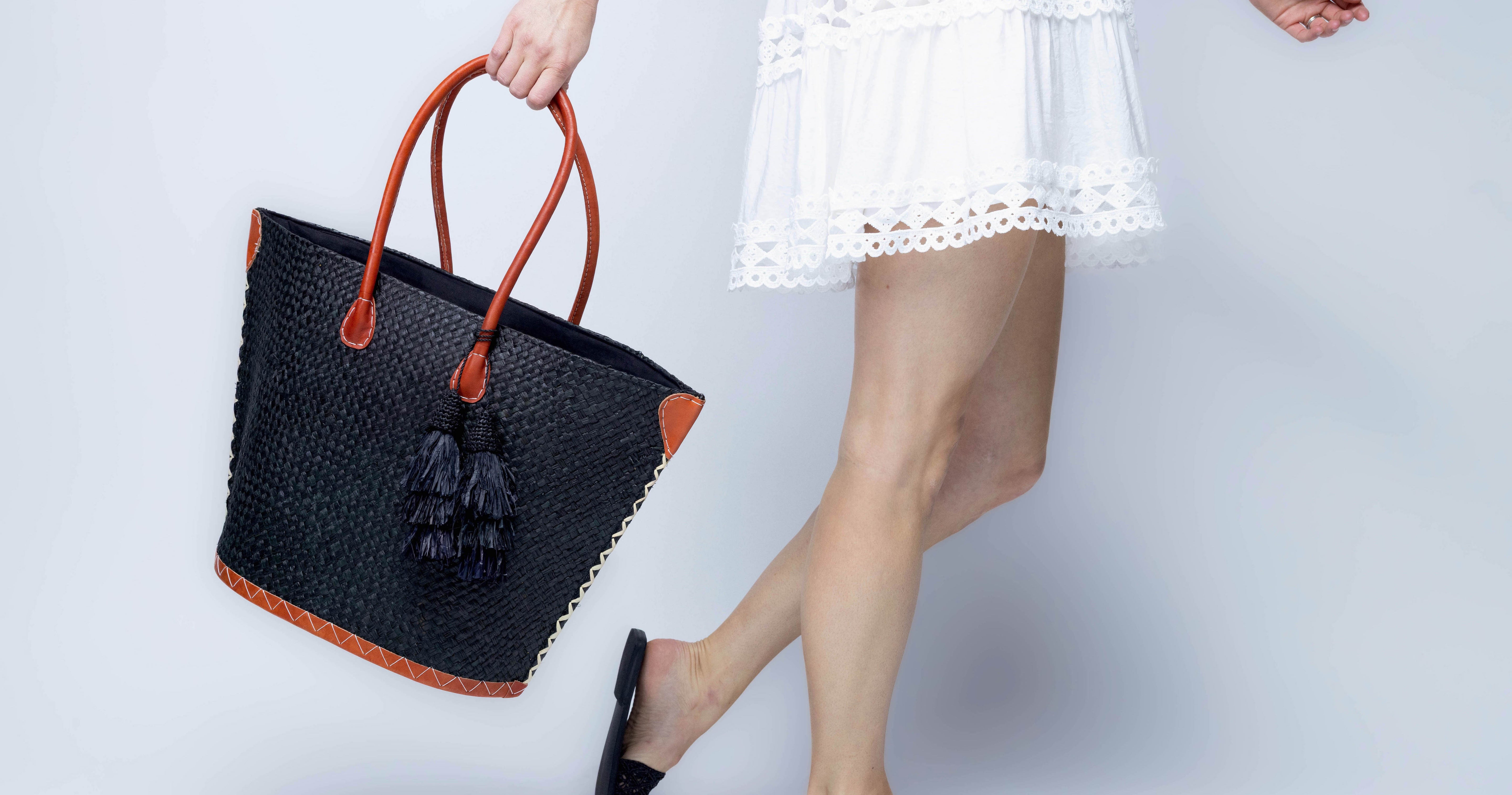 Model wearing Santa Ynez Woven Small Straw Tote Bag with Tassel Charm Embellishment handmade woven raffia palm fiber in a solid hue of black with matching tassel tie charm embellishment and leather handles beach bag - Shebobo
