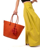 Model wearing Santa Ynez Woven Small Straw Tote Bag with Tassel Charm Embellishment handmade woven raffia palm fiber in a solid hue of coral orange/red with matching tassel tie charm embellishment and leather handles beach bag - Shebobo