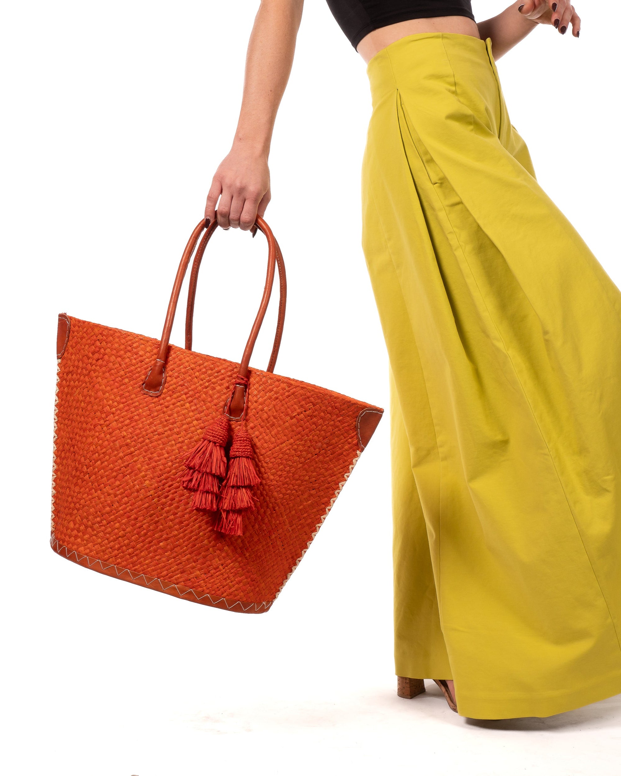 Model wearing Santa Ynez Woven Small Straw Tote Bag with Tassel Charm Embellishment handmade woven raffia palm fiber in a solid hue of coral orange/red with matching tassel tie charm embellishment and leather handles beach bag - Shebobo