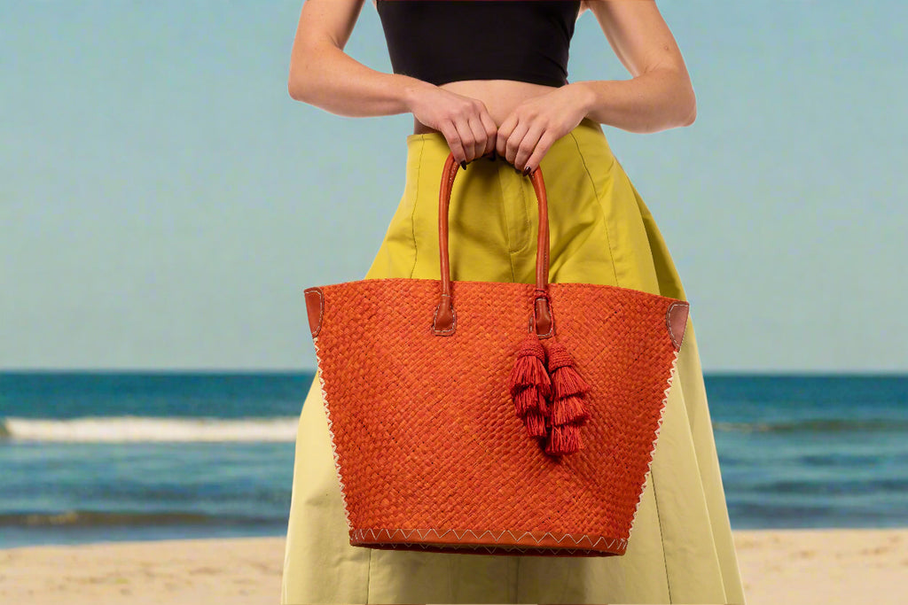 Model wearing Santa Ynez Woven Small Straw Tote Bag with Tassel Charm Embellishment handmade woven raffia palm fiber in a solid hue of coral orange/red with matching tassel tie charm embellishment and leather handles beach bag - Shebobo