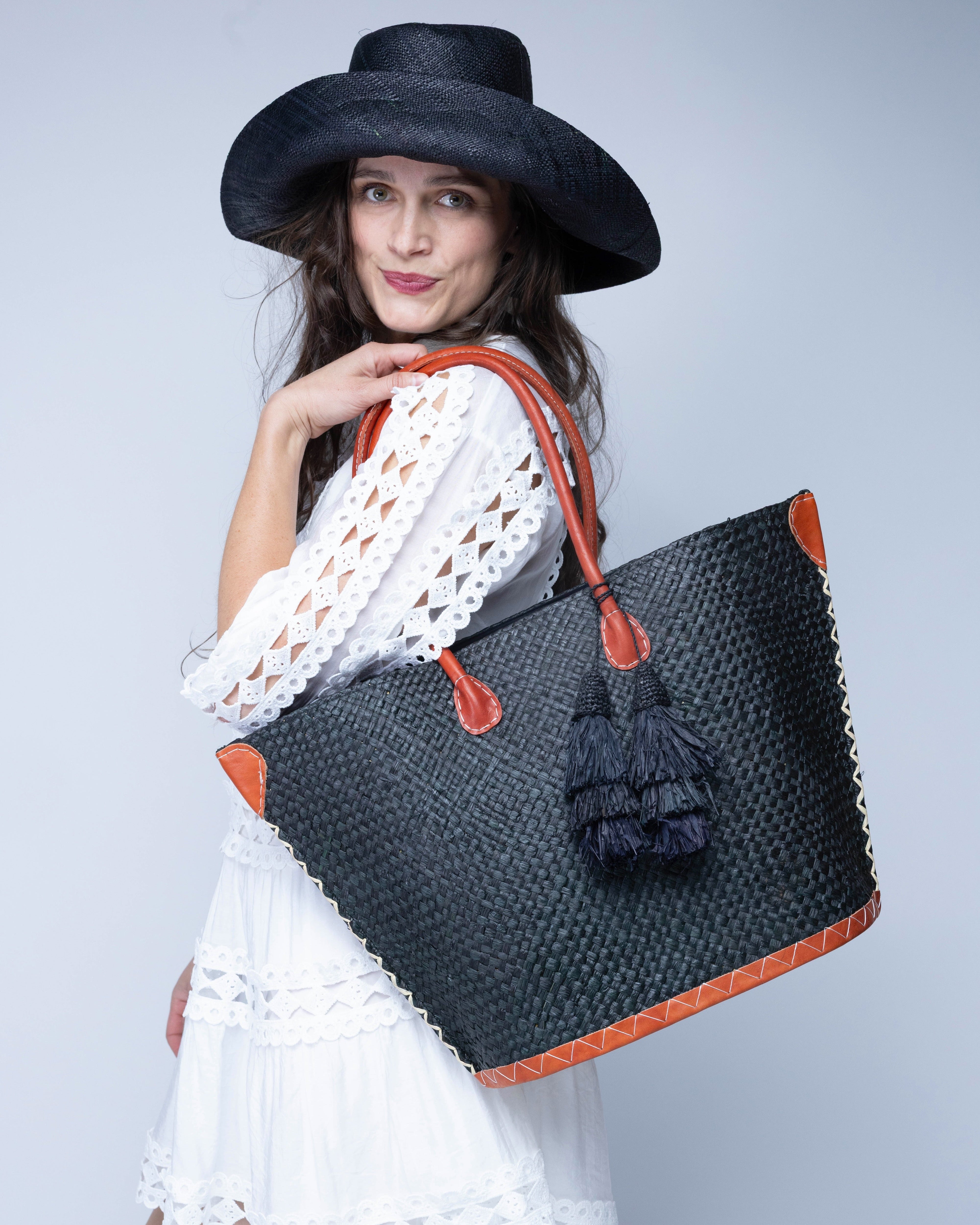 Model wearing Santa Ynez Woven Small Straw Tote Bag with Tassel Charm Embellishment handmade woven raffia palm fiber in a solid hue of black with matching tassel tie charm embellishment and leather handles beach bag - Shebobo (with Monica 5" Wide Brim Packable Straw Sun Hat)