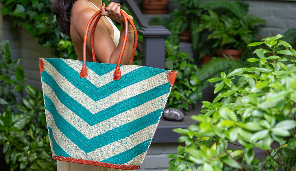 Model wearing Santa Cruz Wide Stripe Chevron Large Straw Tote Bag handmade loomed raffia palm fiber in a two tone diagonal even width bands of color in a wide stripe chevron pattern of teal green/blue and natural straw color on the front and a matching solid hue on the back with leather accents and handles large shoulder bag - Shebobo