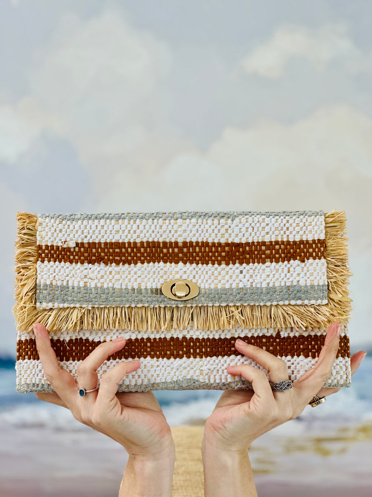 Model wearing Sasha Grey Multi Recycled Cotton Clutch with Raw Fringe Edge handmade woven dyed recycled cotton and natural raffia palm fiber in multicolor horizontal stripe pattern of cinnamon/tobacco/brown, white, and grey with raffia fringe trim edge embellishment - Shebobo