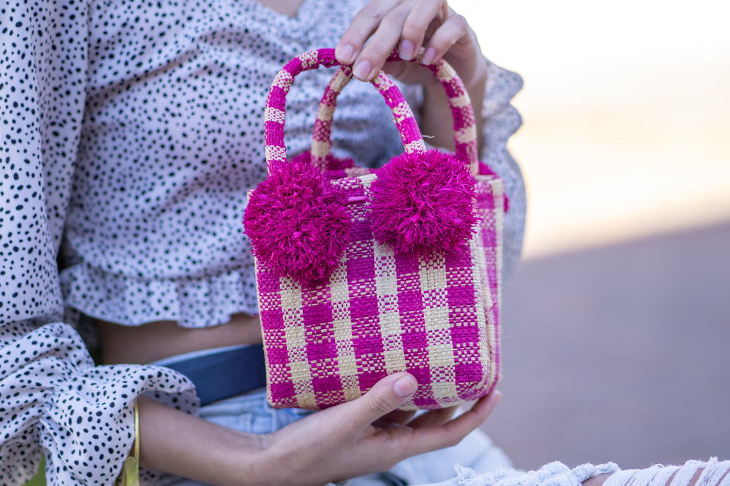 Model wearing Schooner Fuchsia Gingham Straw Mini Bag with Pompom Accent handmade loomed natural raffia palm fiber in two tones of fuchsia pink and natural straw color small gingham plaid pattern with four fuchsia raffia pompom embellishments at the base of each handle attachment tiny purse extra small handbag - Shebobo