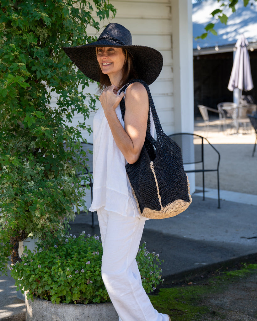 Model wearing Sea See Black Straw Shoulder Bag handmade woven raffia palm fiber in a solid hue of black making a diagonal link pattern with  natural straw color crochet circular bottom and vertical edging along the side panels creating a two tone multicolor handbag/beach bag/purse - Shebobo (with Natasha 7" Wide Brim Woven XL Straw Sun Hats)