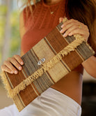 Model wearing Sophie Cinnamon Swirl Multicolor Stripe Straw Clutch with Raw Fringe Edge handmade loomed raffia purse cinnamon/tobacco/light brown, natural straw color, and grey multicolor multi width vertical stripe pattern handbag - Shebobo