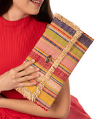Model wearing Sophie Raspberry Swirl Multicolor Stripes Straw Clutch handmade loomed raffia purse handbag  in muti-width vertical bands of pink, yellow, purple, blue, orange, cappuccino light brown, and natural straw color with matching cross stitch edging and natural straw color raw fringe edge envelope shaped bag - Shebobo