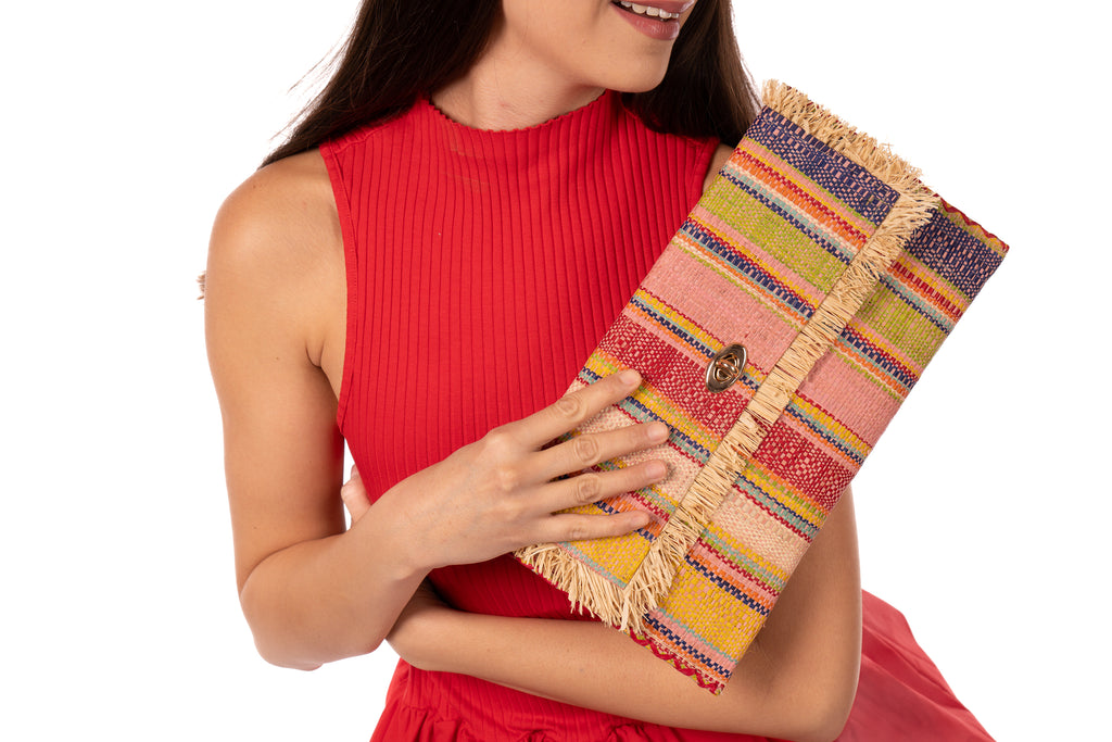 Model wearing Sophie Raspberry Swirl Multicolor Stripes Straw Clutch handmade loomed raffia purse handbag  in muti-width vertical bands of pink, yellow, purple, blue, orange, cappuccino light brown, and natural straw color with matching cross stitch edging and natural straw color raw fringe edge envelope shaped bag - Shebobo