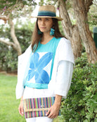 Model wearing Sophie Turquoise Stripes Multicolor Straw Clutch handmade loomed raffia purse handbag with raw fringe edge detailing turquoise, blue, natural, black, red, yellow, purple, orange, green, fuchsia, etc. multi width multicolor vertical stripe pattern envelope shaped bag - Shebobo (with Crochet Fedora - Unisex Straw Hat Natural and Blues woven hat band)