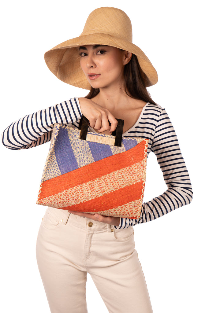 Model wearing Stevie Double Wide Stripe Coral & Denim Square Straw Handbag with Horn Handles handmade loomed raffia palm fiber in even width wide stripe pattern of Coral orange/red and natural straw color oriented on the diagonal of three quarters of the bag with matching stripe pattern of Denim retro/light/blue and natural diagonally on the top left corner quarter shoulder bag handbag - Shebobo (with 5" & 7" Wide Brim Natural Packable Straw Sun Hat)