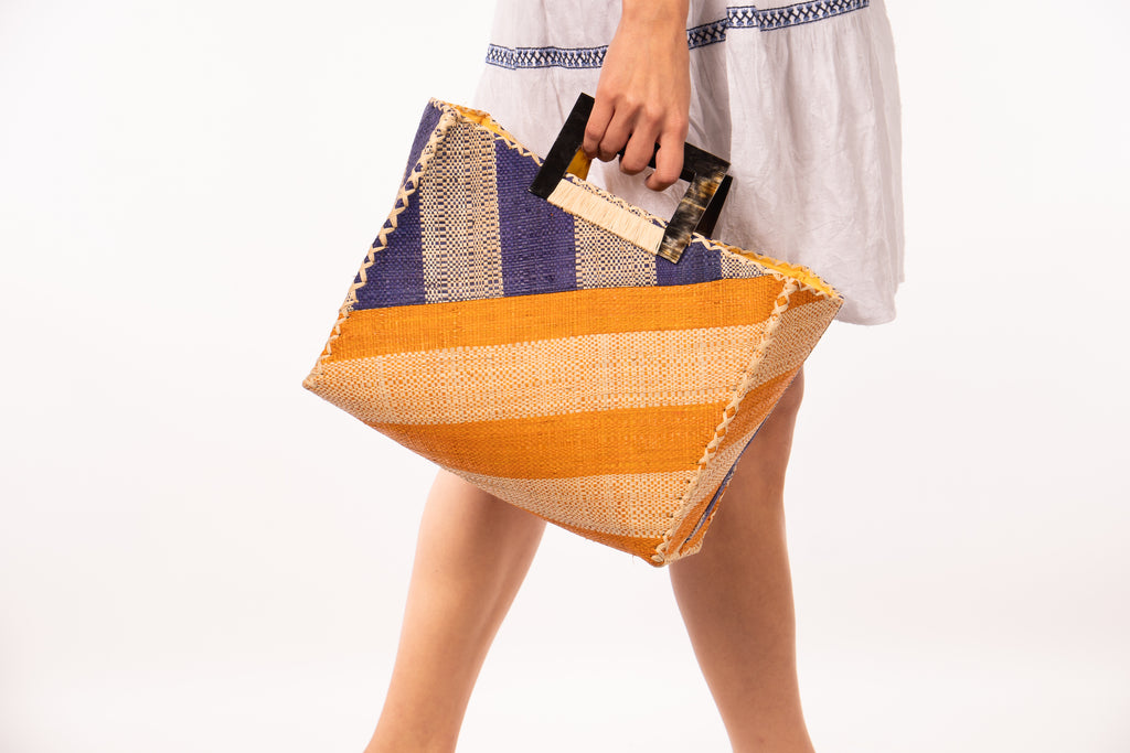 Model wearing Stevie Double Wide Stripe Saffron & Dusk Square Straw Handbag with Horn Handles handmade loomed raffia palm fiber in even width wide stripe pattern of Saffron yellow/orange and natural straw color oriented on the diagonal of three quarters of the bag with matching stripe pattern of Dusk navy/dark/blue and natural diagonally on the top left corner quarter shoulder bag handbag - Shebobo