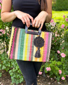 Model wearing Stevie Square Lollipop Multicolor Stripe Straw Handbag with Horn Handles handmade loomed raffia with yellow, saffron, light blue, green, red, black, fuchsia, orange, natural, etc. varying width vertical stripe pattern purse with matching black liner, cross stitch edging, and handle attachment - Shebobo