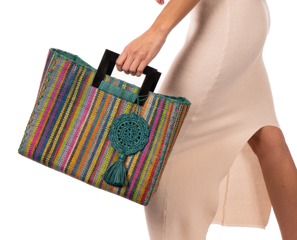 Model wearing Stevie Square Turquoise Stripes Multicolor Straw Handbag with Horn Handles handmade loomed raffia with turquoise, navy, light blue, purple, red, black, fuchsia, orange, yellow, lime, etc. varying width vertical stripe pattern purse with matching turquoise liner, cross stitch edging, handle attachment, and crochet doily tassel embellishment - Shebobo