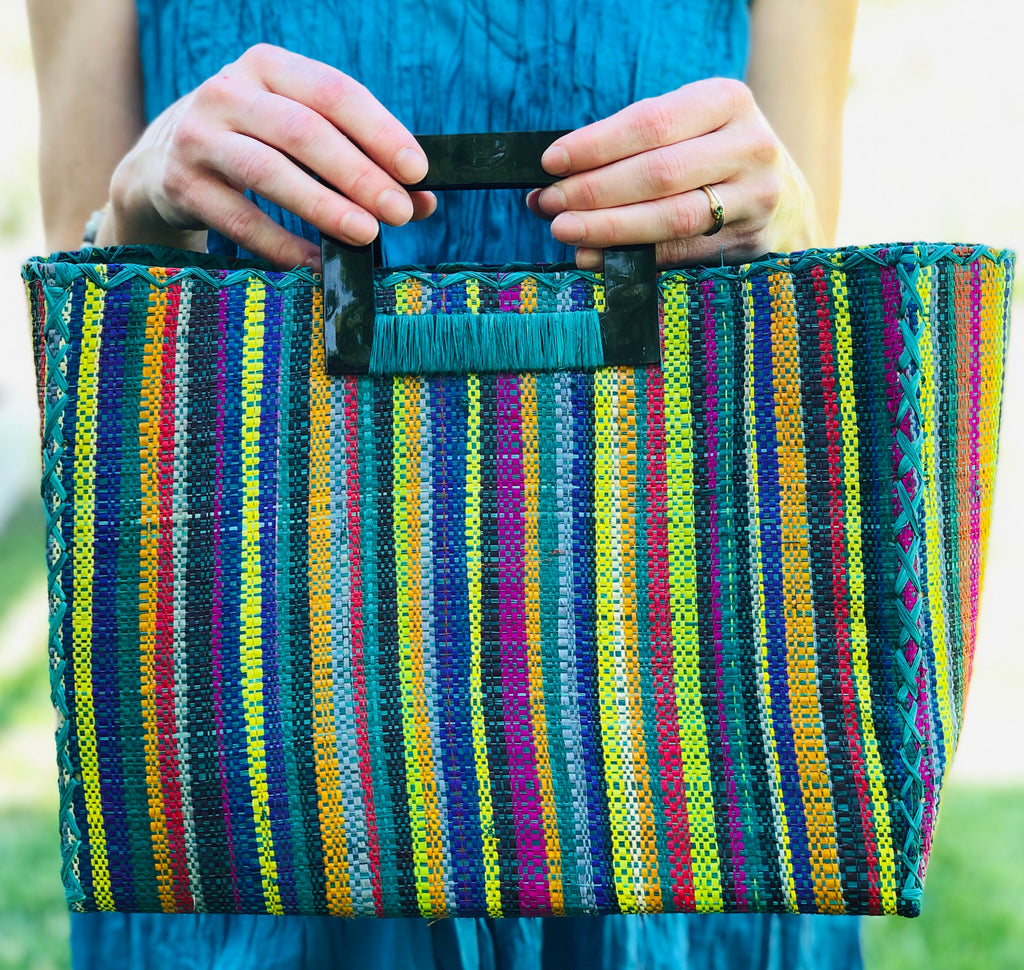 Model wearing Stevie Square Turquoise Stripes Multicolor Straw Handbag with Horn Handles handmade loomed raffia with turquoise, navy, light blue, purple, red, black, fuchsia, orange, yellow, lime, etc. varying width vertical stripe pattern purse with matching turquoise liner, cross stitch edging, and handle attachment - Shebobo