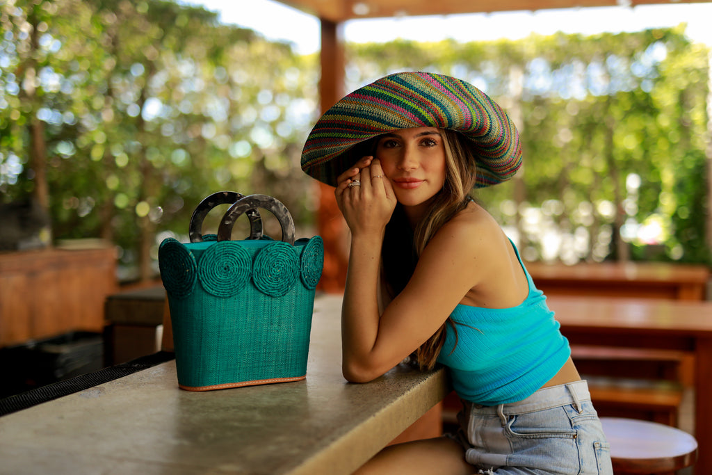Model wearing Tazi Disc Straw Handbag with Horn Handle handmade loomed raffia purse in turquoise blue with wrapped raffia disc embellishment small bag - Shebobo (with 5" Turquoise Stripes Multicolor Packable Straw Sun Hat)
