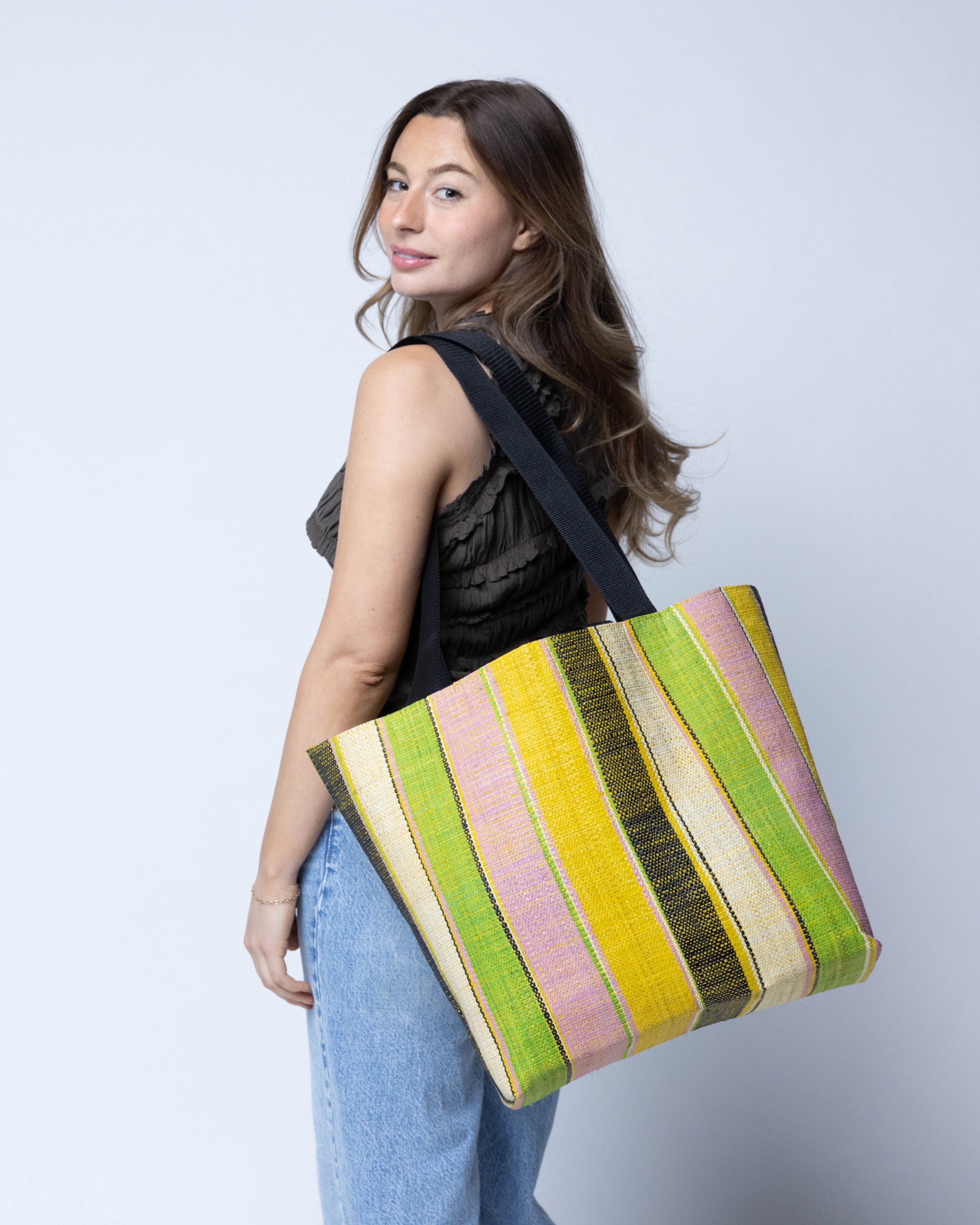 Model wearing Tiburon Lemonade Swirl Straw Duffle Bag Shoulder & Handbag Combination handmade loomed natural raffia palm fiber in a multicolor vertical stripe pattern of multi width bands of black, saffron yellow, lime green, purple pink, and natural straw color, etc. with four black webbing handles sports tote bag purse - Shebobo