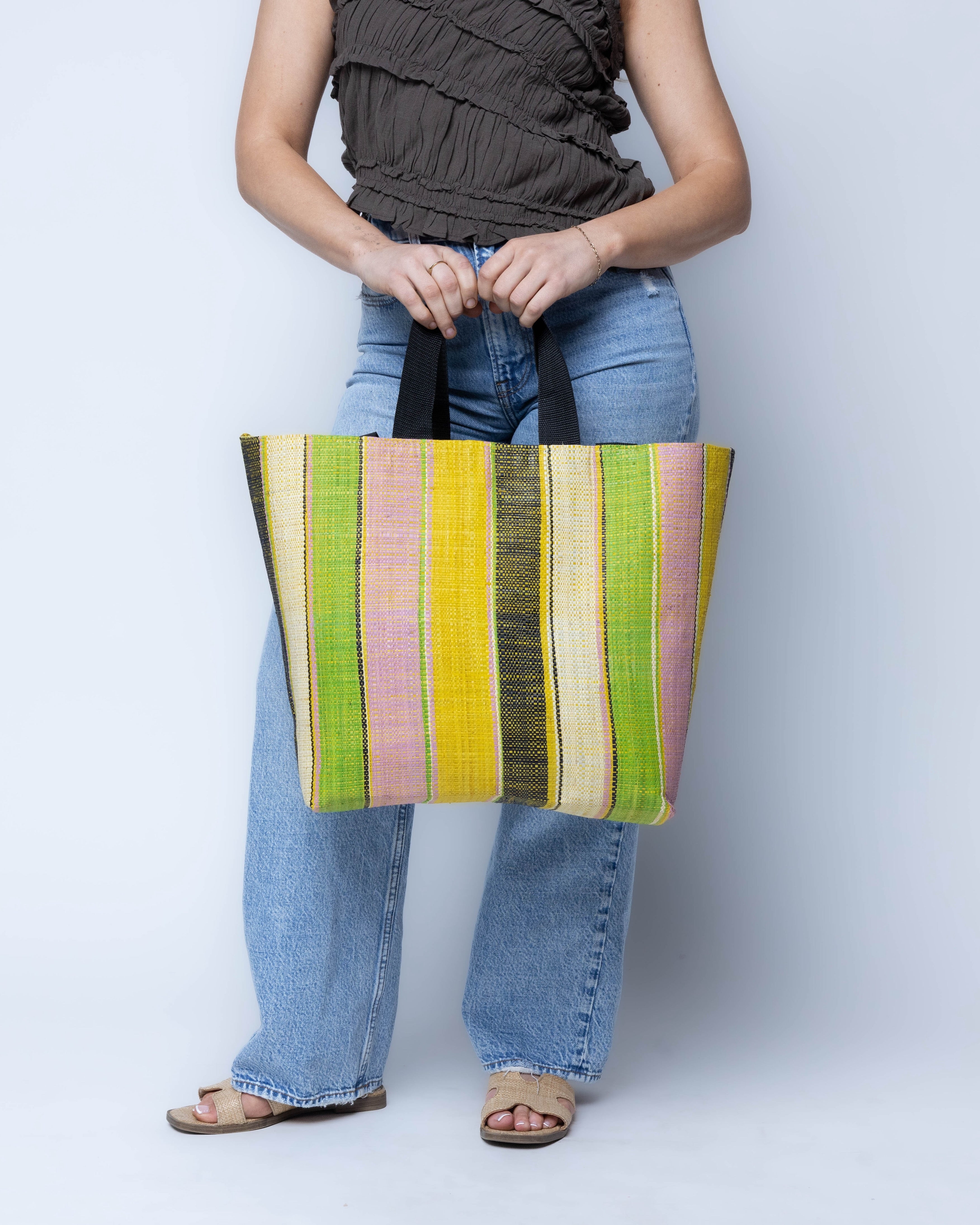 Model wearing Tiburon Lemonade Swirl Straw Duffle Bag Shoulder & Handbag Combination handmade loomed natural raffia palm fiber in a multicolor vertical stripe pattern of multi width bands of black, saffron yellow, lime green, purple pink, and natural straw color, etc. with four black webbing handles sports tote bag purse - Shebobo