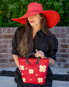 Model wearing Tulum Straw Bucket Bag with Flower Embroidery and Horn Handle Red Purse with Ruffle Edge handmade loomed raffia palm fiber in a solid hue of red with natural straw color hand embroidered floral pattern with multiple sized flowers and raffia ruffle on the opening  handbag - Shebobo (with 7" Wide Brim Natasha Red Woven XL Straw Sun Hats)