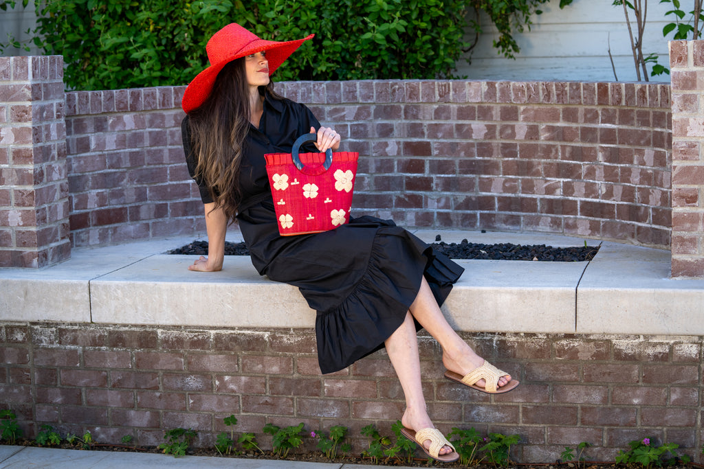 Model wearing Tulum Straw Bucket Bag with Flower Embroidery and Horn Handle Red Purse with Ruffle Edge handmade loomed raffia palm fiber in a solid hue of red with natural straw color hand embroidered floral pattern with multiple sized flowers and raffia ruffle on the opening  handbag - Shebobo (with 7" Wide Brim Natasha Red Woven XL Straw Sun Hats)