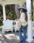 Model wearing Valencia Mini Crochet Petit Straw Basket Handbag with Flower Embellishment handmade natural raffia palm fiber in a solid hue of Natural straw color crochet in bands of horizontal link pattern texture with integrated strap handle and large flower embellishment - Shebobo (with Monica 5" & 7" Wide Brim Solid Color Packable Straw Sun Hats)