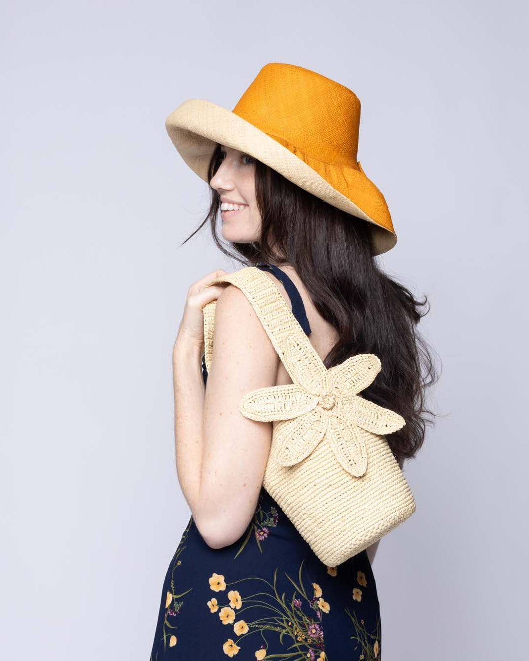 Model wearing Valencia Mini Crochet Petit Straw Basket Handbag with Flower Embellishment handmade natural raffia palm fiber in a solid hue of Natural straw color crochet in bands of horizontal link pattern texture with integrated strap handle and large flower embellishment - Shebobo