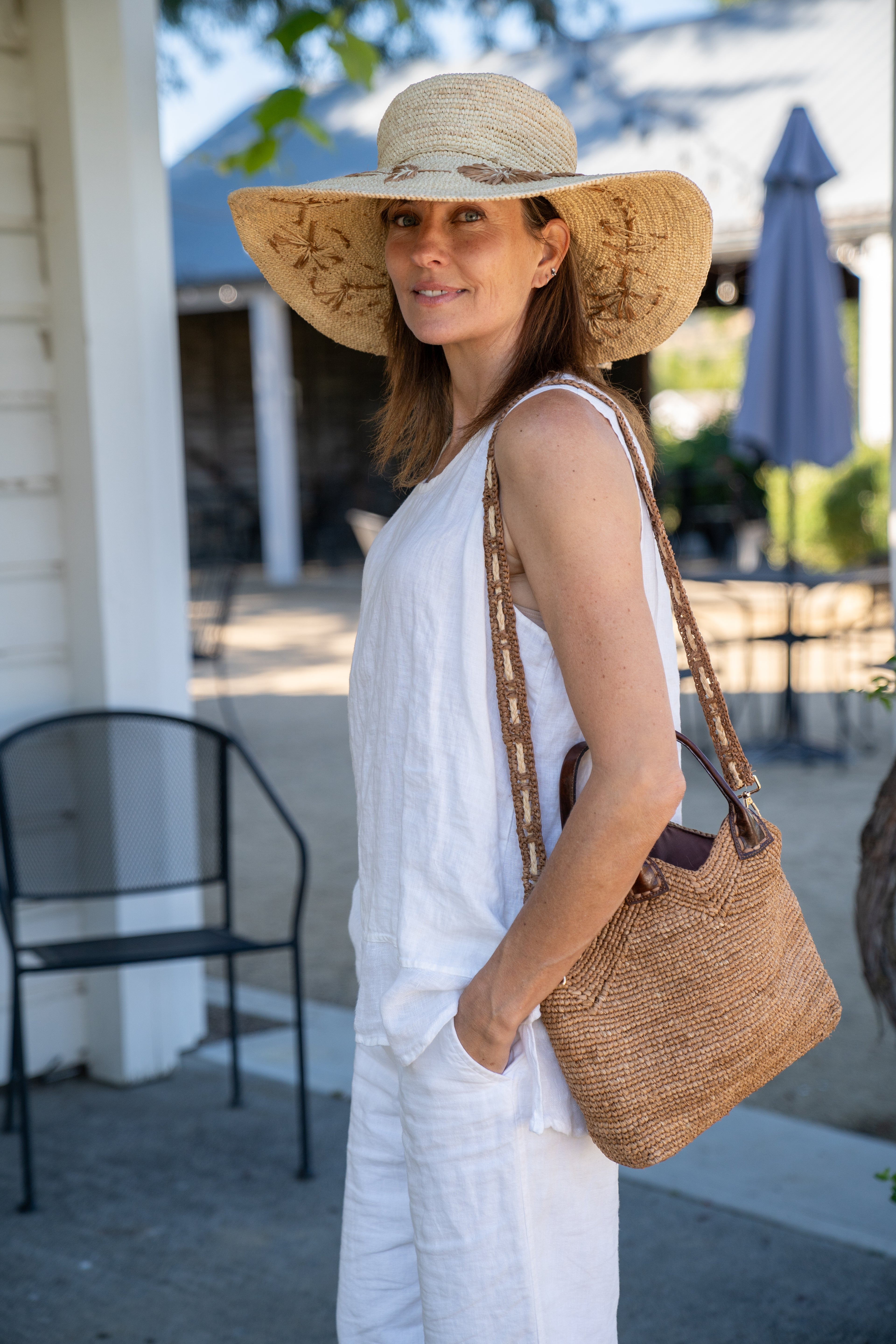 Model wearing Victoria Crochet Straw Handbag Convertible Crossbody Bag handmade crochet natural raffia palm fiber in a solid hue of cappuccino tea/light/brown in a subtle texture of horizontal link bands pattern across the bag and m shape around the top of the bag with dark brown leather handles and removable two tone natural and cappuccino tea/light/brown crossbody strap convertible purse - Shebobo (with Gigi 5.5" Brim Floral Embroidered Crochet Straw Sun Hats)