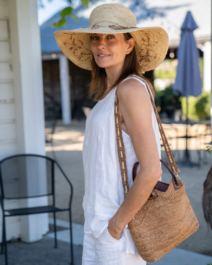 Model wearing Victoria Crochet Straw Handbag Convertible Crossbody Bag handmade crochet natural raffia palm fiber in a solid hue of cappuccino tea/light/brown in a subtle texture of horizontal link bands pattern across the bag and m shape around the top of the bag with dark brown leather handles and removable two tone natural and cappuccino tea/light/brown crossbody strap convertible purse - Shebobo (with Gigi 5.5" Brim Floral Embroidered Crochet Straw Sun Hats)