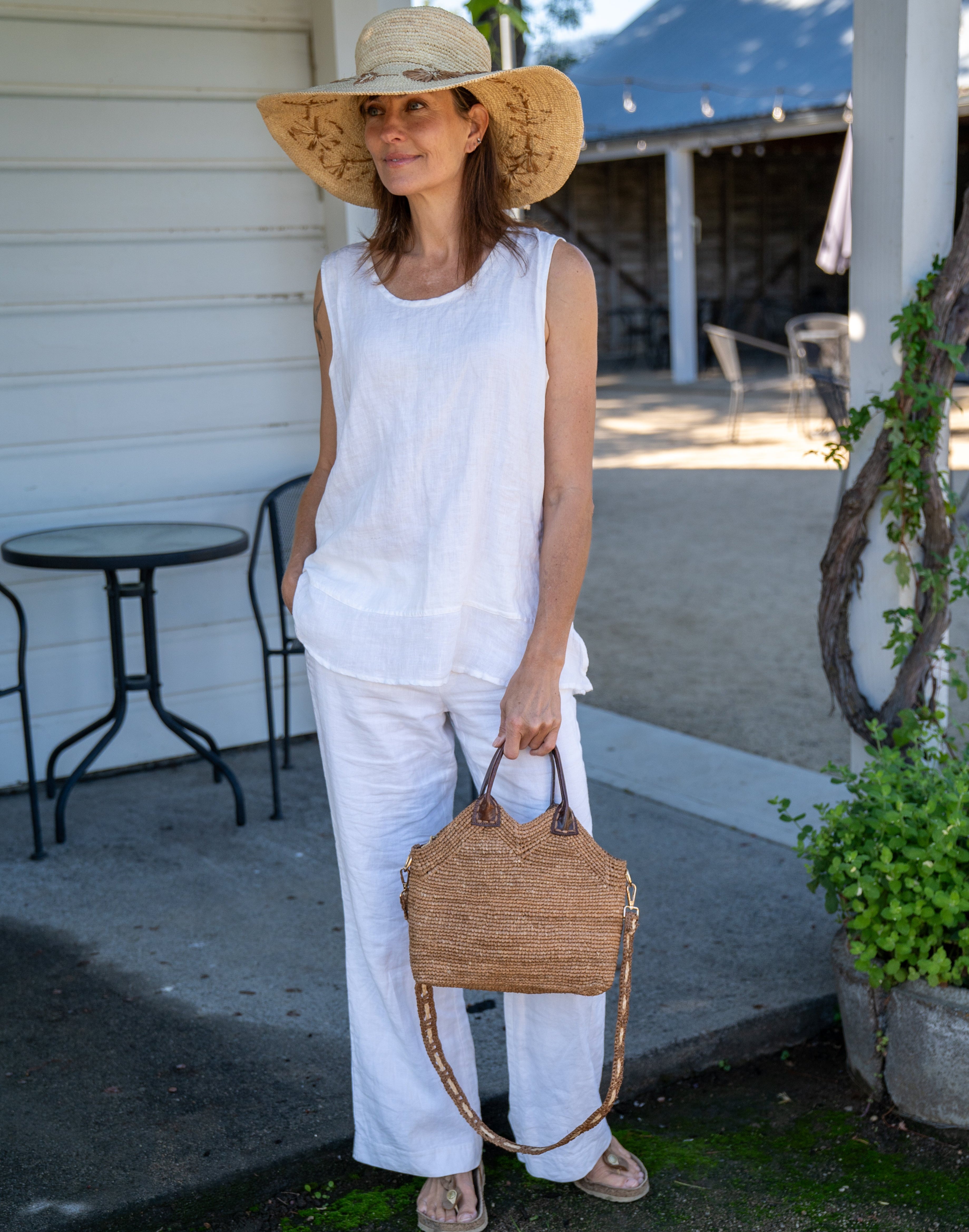 Model wearing Victoria Crochet Straw Handbag Convertible Crossbody Bag handmade crochet natural raffia palm fiber in a solid hue of cappuccino tea/light/brown in a subtle texture of horizontal link bands pattern across the bag and m shape around the top of the bag with dark brown leather handles and removable two tone natural and cappuccino tea/light/brown crossbody strap convertible purse - Shebobo (with Gigi 5.5" Brim Floral Embroidered Crochet Straw Sun Hats)