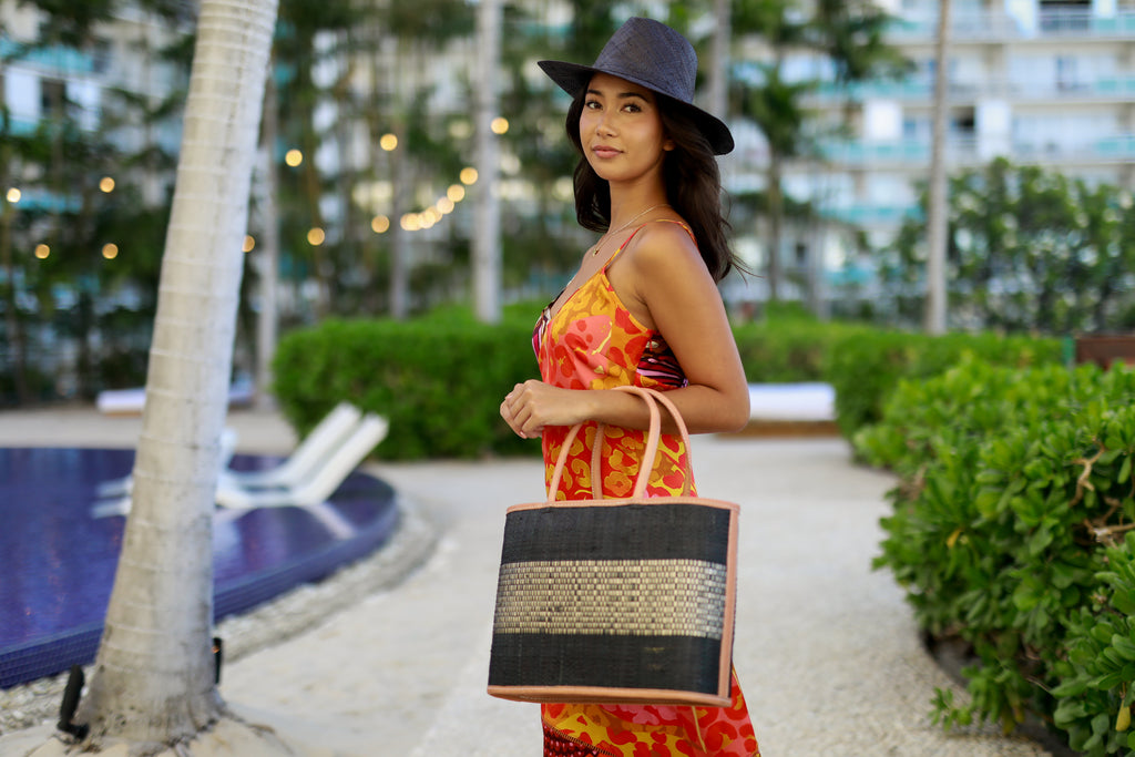 Model wearing Wynwood Black Straw Basket Bag Handbag with Metallic Detailing handmade loomed raffia in black and silver metallic vegan leather in three evenly sized horizontal bands of color with the metallic weave centered on the purse with leather binding and handles - Shebobo (with Panama Black Unisex Straw Hat)