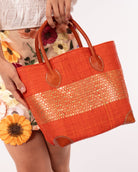 Model wearing Wynwood Coral Straw Basket Bag with Metallic Detailing Handmade loomed raffia palm fiber in a solid hue of coral orange/red with a wide horizontal band of metallic strands woven in seamlessly handbag purse with leather handles - Shebobo