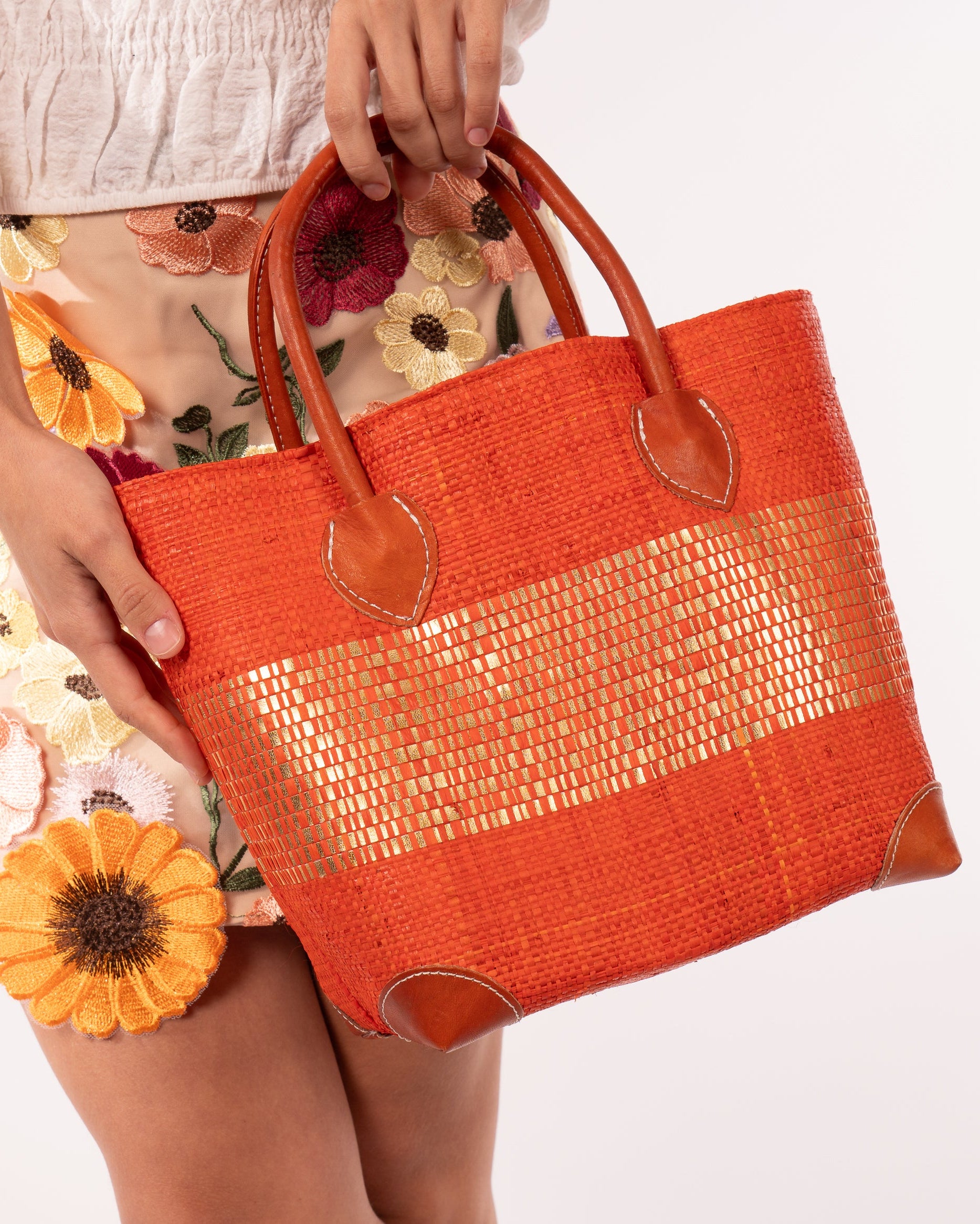 Model wearing Wynwood Coral Straw Basket Bag with Metallic Detailing Handmade loomed raffia palm fiber in a solid hue of coral orange/red with a wide horizontal band of metallic strands woven in seamlessly handbag purse with leather handles - Shebobo
