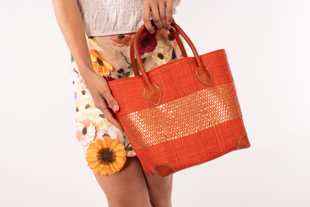 Model wearing Wynwood Coral Straw Basket Bag with Metallic Detailing Handmade loomed raffia palm fiber in a solid hue of coral orange/red with a wide horizontal band of metallic strands woven in seamlessly handbag purse with leather handles - Shebobo