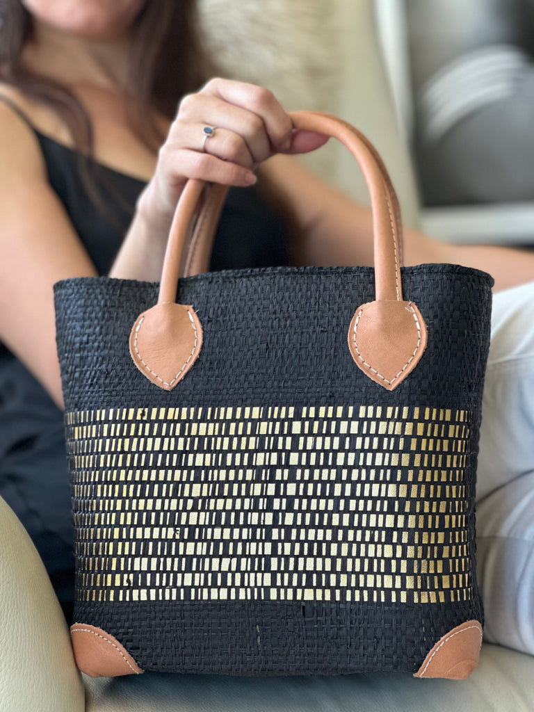Model wearing Wynwood Black Straw Basket Bag with Metallic Detailing Handmade loomed raffia palm fiber in a solid hue of black with a wide horizontal band of metallic strands woven in seamlessly handbag purse with leather handles - Shebobo