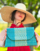 Model wearing Wynwood Turquoise Straw Handbag with Metallic Detailing & Horn Handle handmade loomed raffia in turquoise blue and metallic vegan leather in three vertical bands of color with the metallic weave centered on the purse under the handles with 7" Wide Brim Natasha Natural Woven Straw Sun Hat - Shebobo
