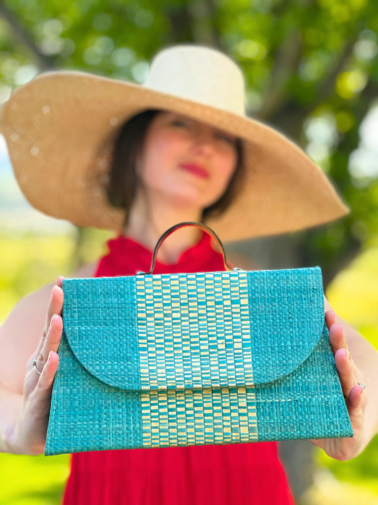 Model wearing Wynwood Turquoise Straw Handbag with Metallic Detailing & Horn Handle handmade loomed raffia in turquoise blue and metallic vegan leather in three vertical bands of color with the metallic weave centered on the purse under the handles with 7" Wide Brim Natasha Natural Woven Straw Sun Hat - Shebobo