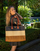 Model wearing Zafran 3 Tone Color Block Beach Straw Bag with Plastic Liner handmade loomed raffia palm fiber in three equally sized horizontal bands of black, natural straw color, and cinnamon/tobacco/dark brown with wood button and leather handles - Shebobo