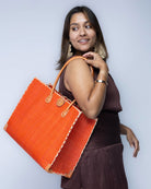 Model wearing Zafran Straw Beach Bag with Plastic Liner handmade loomed raffia in a solid hue of Coral orange/red with contrasting natural straw color cross stitch edge binding, wood button closure, and leather handles & feet plus assorted print plastic lining - Shebobo