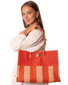 Model wearing Zafran Two Tone Wide Stripe Coral and Natural Straw Beach Bag handmade loomed raffia palm fiber in a solid hue of coral orange/red on the top half of the bag and a two tone even width vertical wide stripe pattern of coral and natural straw color on the bottom half of the bag with leather handles and feet and matching crosshatch stitch edging shoulder bag - Shebobo