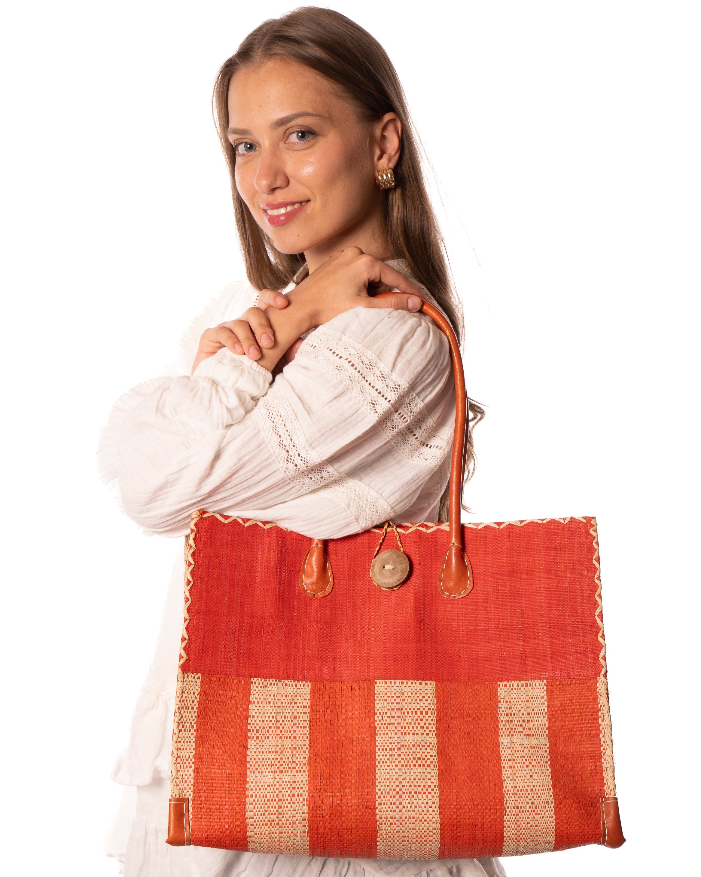 Model wearing Zafran Two Tone Wide Stripe Coral and Natural Straw Beach Bag handmade loomed raffia palm fiber in a solid hue of coral orange/red on the top half of the bag and a two tone even width vertical wide stripe pattern of coral and natural straw color on the bottom half of the bag with leather handles and feet and matching crosshatch stitch edging shoulder bag - Shebobo