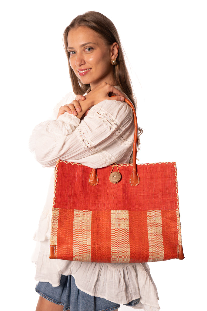 Model wearing Zafran Two Tone Wide Stripe Coral and Natural Straw Beach Bag handmade loomed raffia palm fiber in a solid hue of coral orange/red on the top half of the bag and a two tone even width vertical wide stripe pattern of coral and natural straw color on the bottom half of the bag with leather handles and feet and matching crosshatch stitch edging shoulder bag - Shebobo