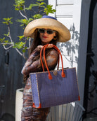 Model wearing Zafran Straw Beach Bag with Plastic Liner handmade loomed raffia in a solid hue of Denim retro/light/blue with contrasting natural straw color cross stitch edge binding, wood button closure, and leather handles & feet plus assorted print plastic lining - Shebobo (with 7" Wide Brim Two Tone Packable Straw Sun Hat)