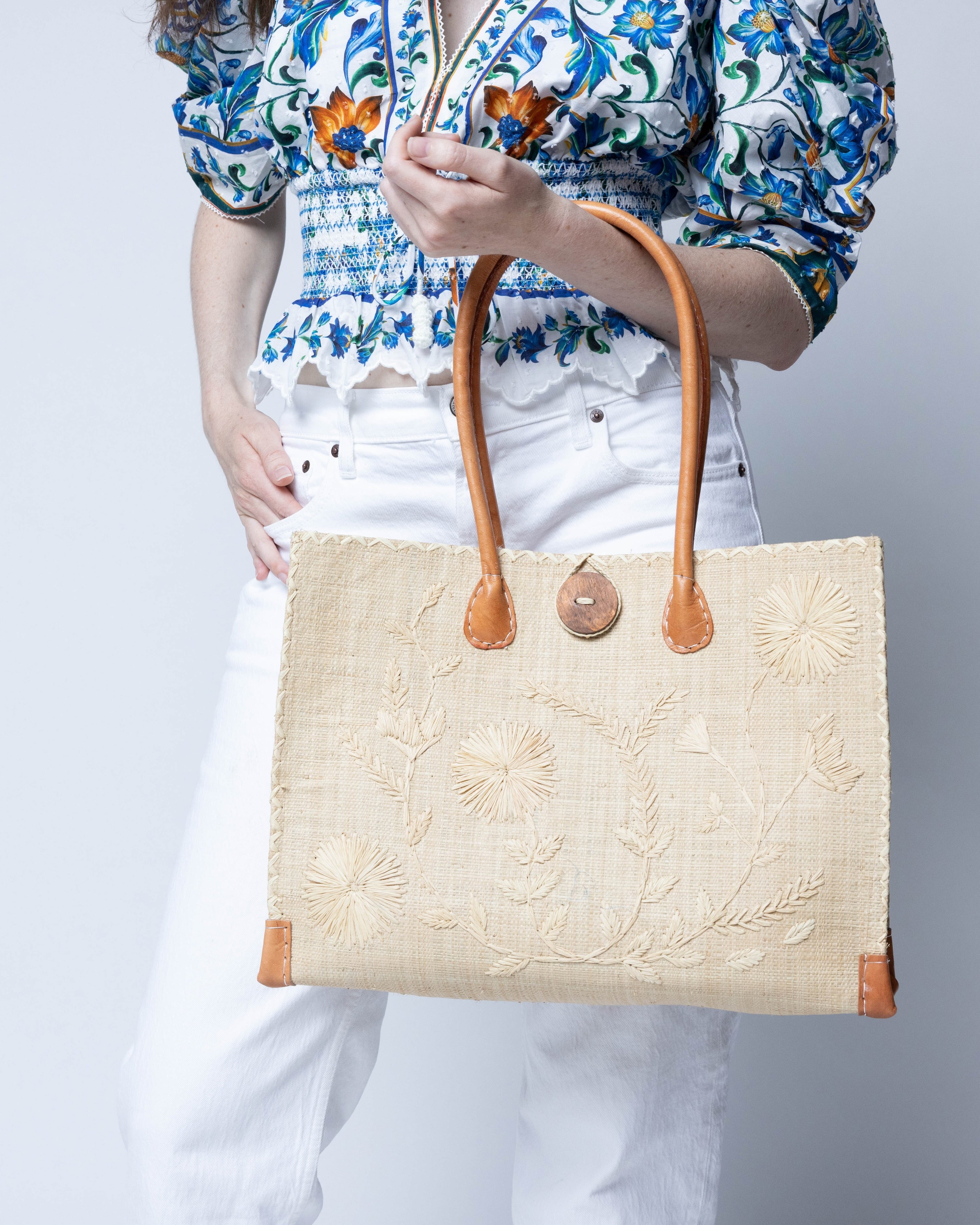 Model wearing Zafran Natural Embroidered Straw Beach Bag with Floral Pattern handmade natural raffia palm fiber in a solid hue of natural straw color with asymmetrical floral leaf, vine, and flower pattern in Natural with matching cross-stitch edging, leather handles, and wooden button closure shoulder bag handbag tote - Shebobo