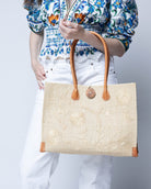 Model wearing Zafran Natural Embroidered Straw Beach Bag with Floral Pattern handmade natural raffia palm fiber in a solid hue of natural straw color with asymmetrical floral leaf, vine, and flower pattern in Natural with matching cross-stitch edging, leather handles, and wooden button closure shoulder bag handbag tote - Shebobo
